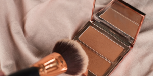 Bronzer e contorno principais diferenças. Foto embalagem de bronzer sobre a cama com lençol branco e pincel dourado.