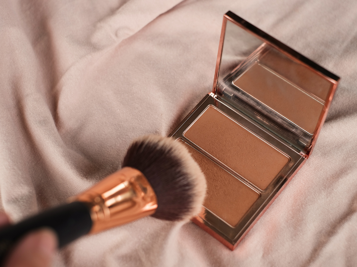 Bronzer e contorno principais diferenças. Foto embalagem de bronzer sobre a cama com lençol branco e pincel dourado. 