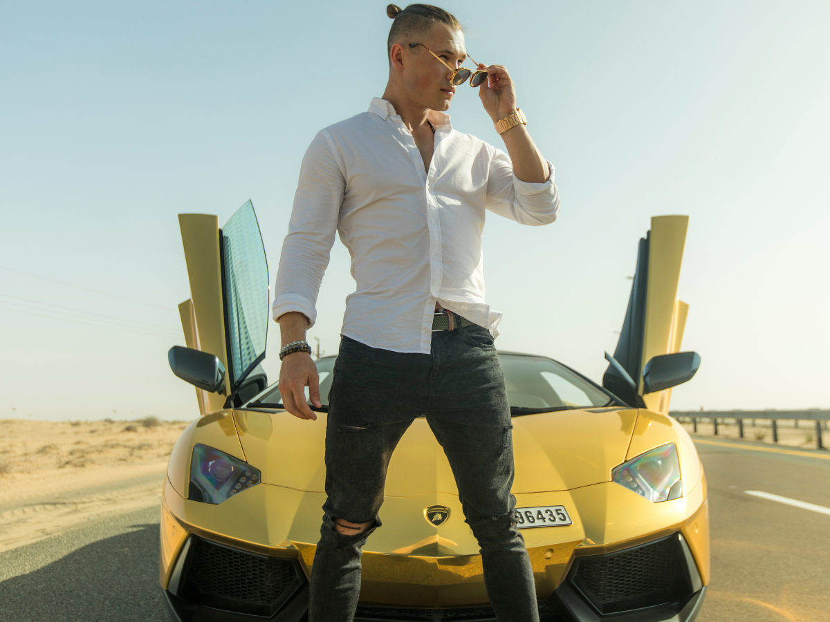 Quiet Luxury. Foto homem branco com camisa branca e calça jeans furada no joelho em frente a um carro de luxo