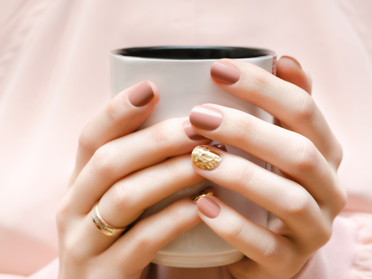 Nail Arts elegantes para mulheres de 30 anos. Foto aparece mãos segurando uma xícara branca. Unhas trazem esmalte nude e detalhes em dourado.