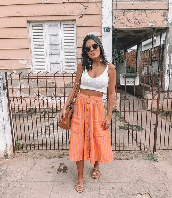 Looks para usar com rasteirinha. Foto traz mulher com pele morena, de óculos escuros pretos, vestida com cropped branco e saia midi de cor laranja com botões. Nos pés ela traz uma rasteirinha de cordas de cor nude.