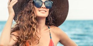 Cabelo no verão. Foto traz uma mulher sorrindo com cabelos cacheados e protegidos com um chapéu e óculos escuros