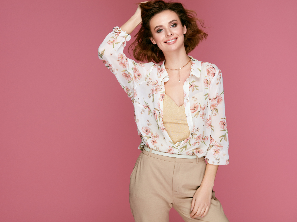 Roupas para o Escritório. Foto traz mulher sorrindo e fundo rosa. Sua roupa traz camiseta soltinha de cor branca com estampas de flores em rosa. Calça bege