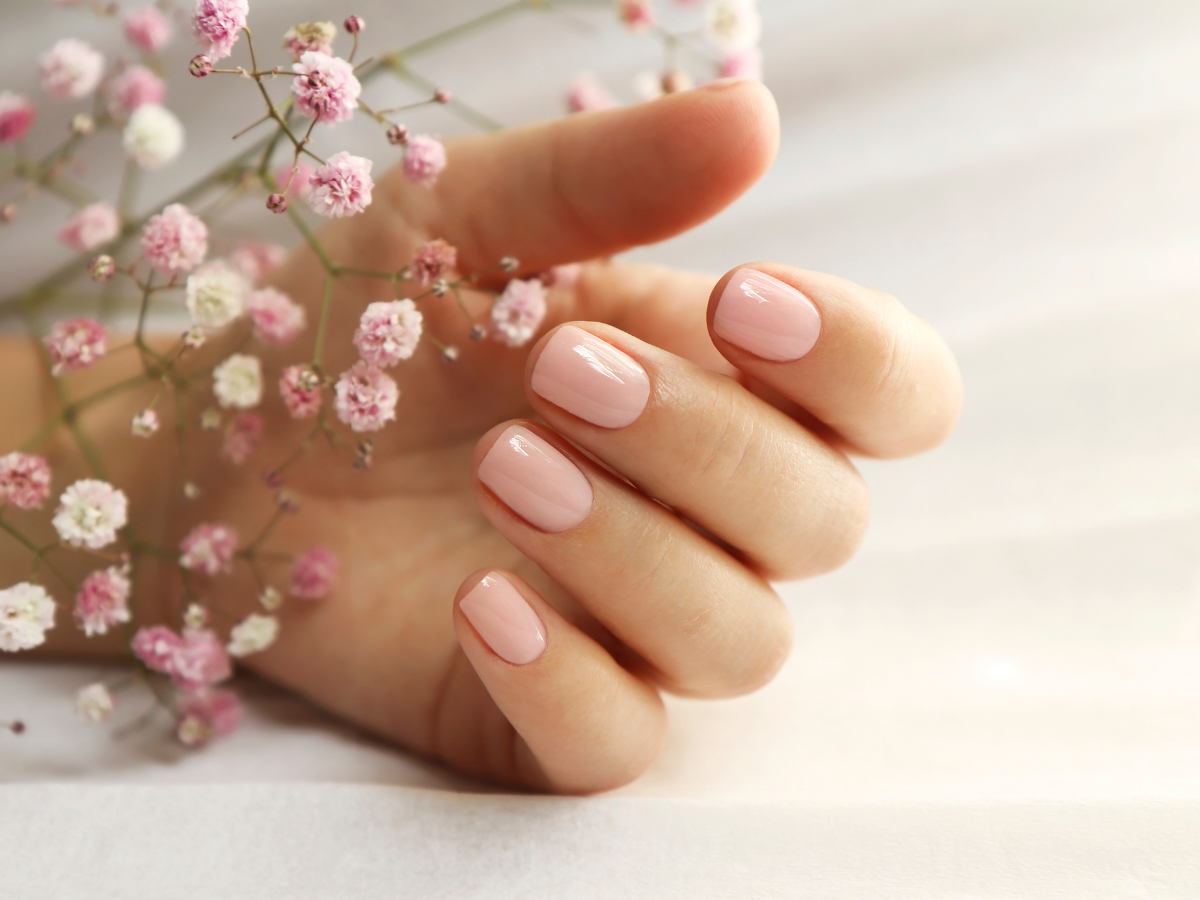 unhas curtas delicadas. Foto mostra mão sobre flores e com a unha pintada de nude